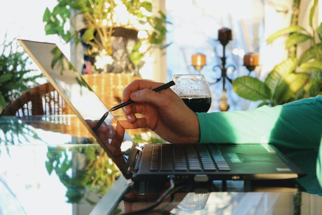 Person using a stylus on a reflective laptop screen in a sunlit room with plants and a glass of wine nearby