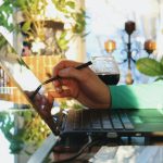 Person using a stylus on a reflective laptop screen in a sunlit room with plants and a glass of wine nearby