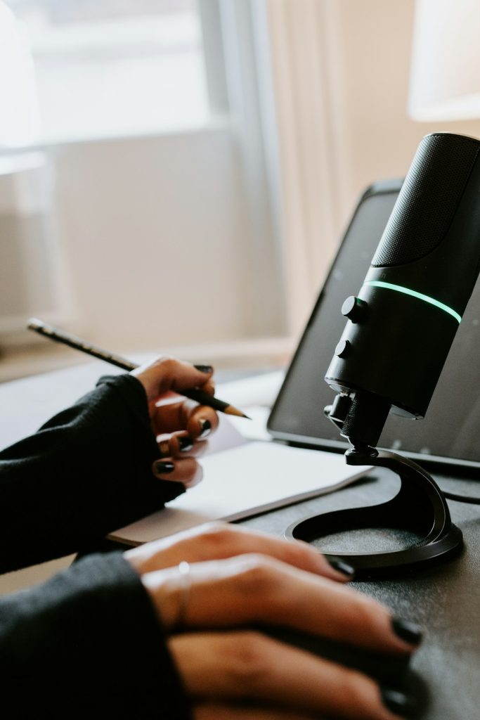 Person writing notes with pencil beside a microphone and tablet, focused on podcast planning or recording session